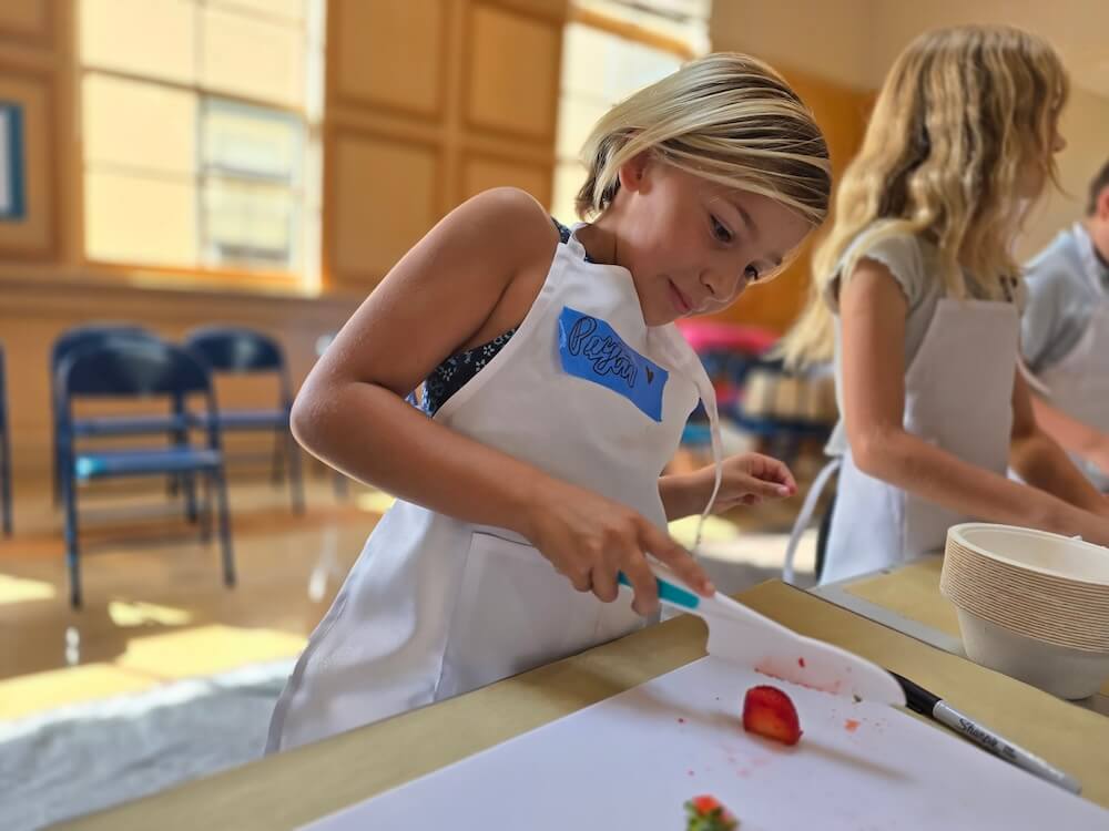 A young girl in an apron carefully cuts strawberries on a cutting board, focused on her task in a bright kitchen.