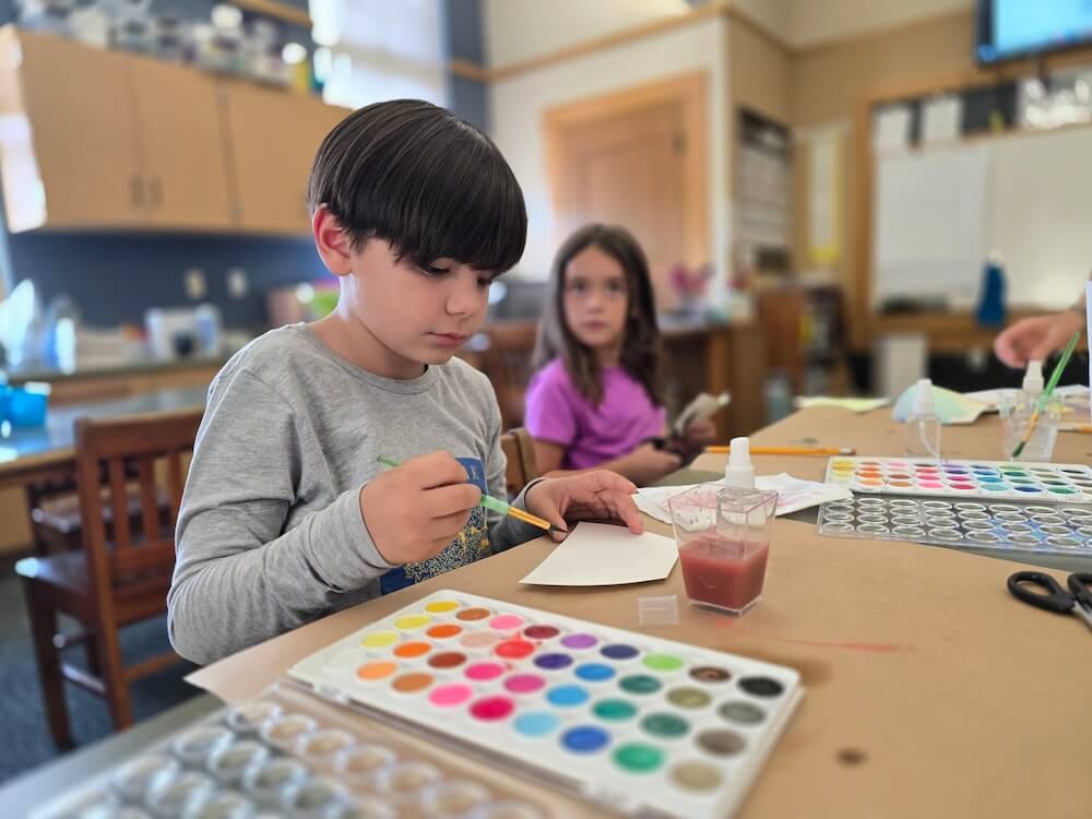 A boy and girl collaborate on a project together at their classroom desk, surrounded by books and school supplies.