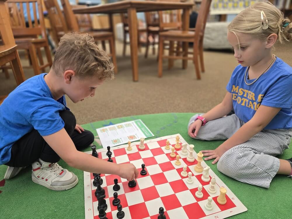 Two children engaged in a chess game at a classroom table, focused on their strategy and the chessboard.
